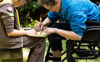 Woman in wheelchair helps girl in Brownie uniform sitting on bench with crafting