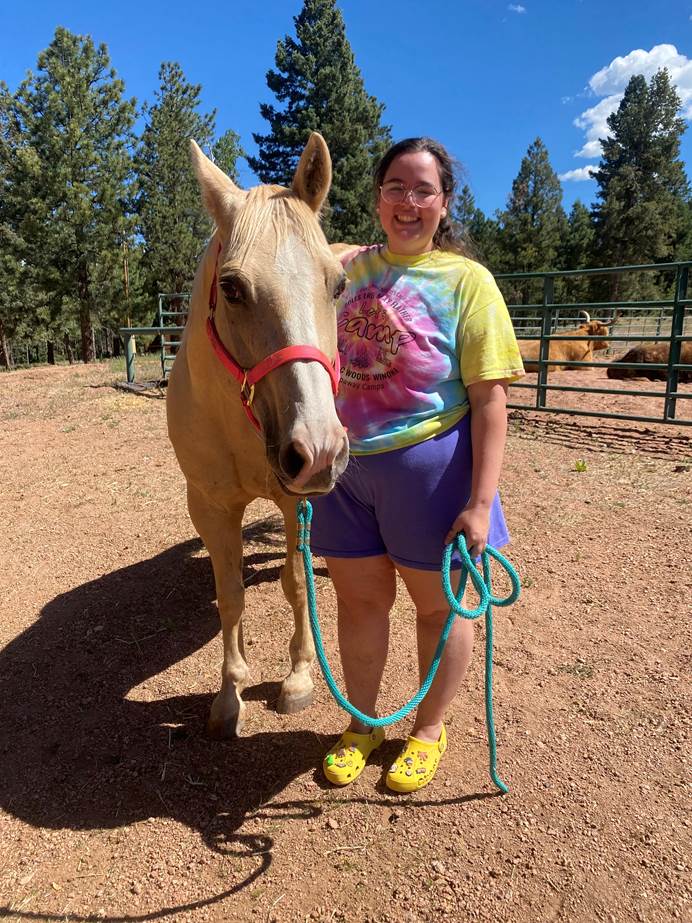 A woman with brown hair in a ponytail, wearing a brightly coloured t-shirt and shorts stands next to a horse smiling at the camera