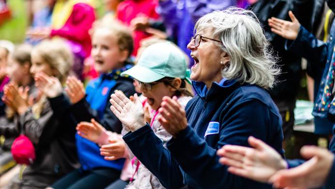 A volunteer is sitting outdoors, clapping and cheering, surrounded by other girls and volunteers