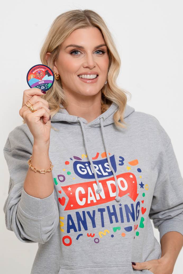 Ashley James is standing, holding the International Women's Day woven badge. She is wearing a hoodie that says girls can do anything