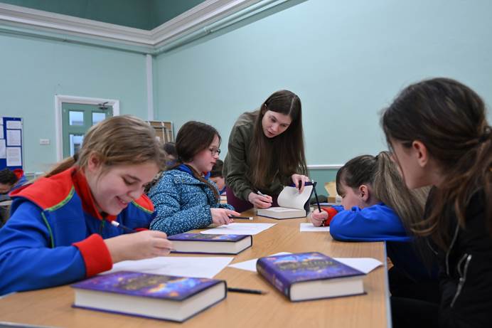 Four Guides in uniform are sitting at a table doing an activity on paper. Author Philippa Leathley is standing up, leaning over to talk to the girls